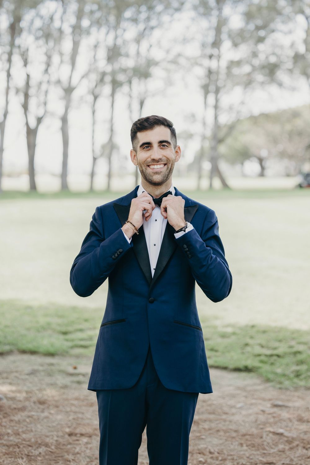 A person in a navy suit makes playful gestures while standing in a grassy outdoor setting.