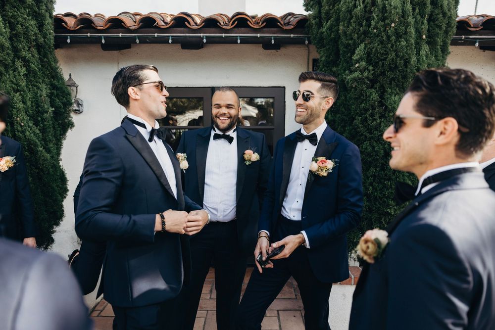 Wedding party members in dark suits socialize in an outdoor courtyard setting.