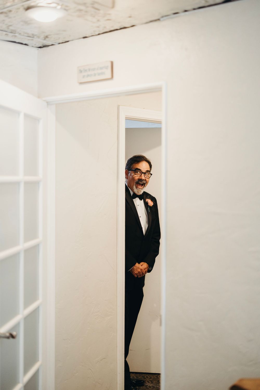 A person in formal attire peeks playfully from behind a white doorway.