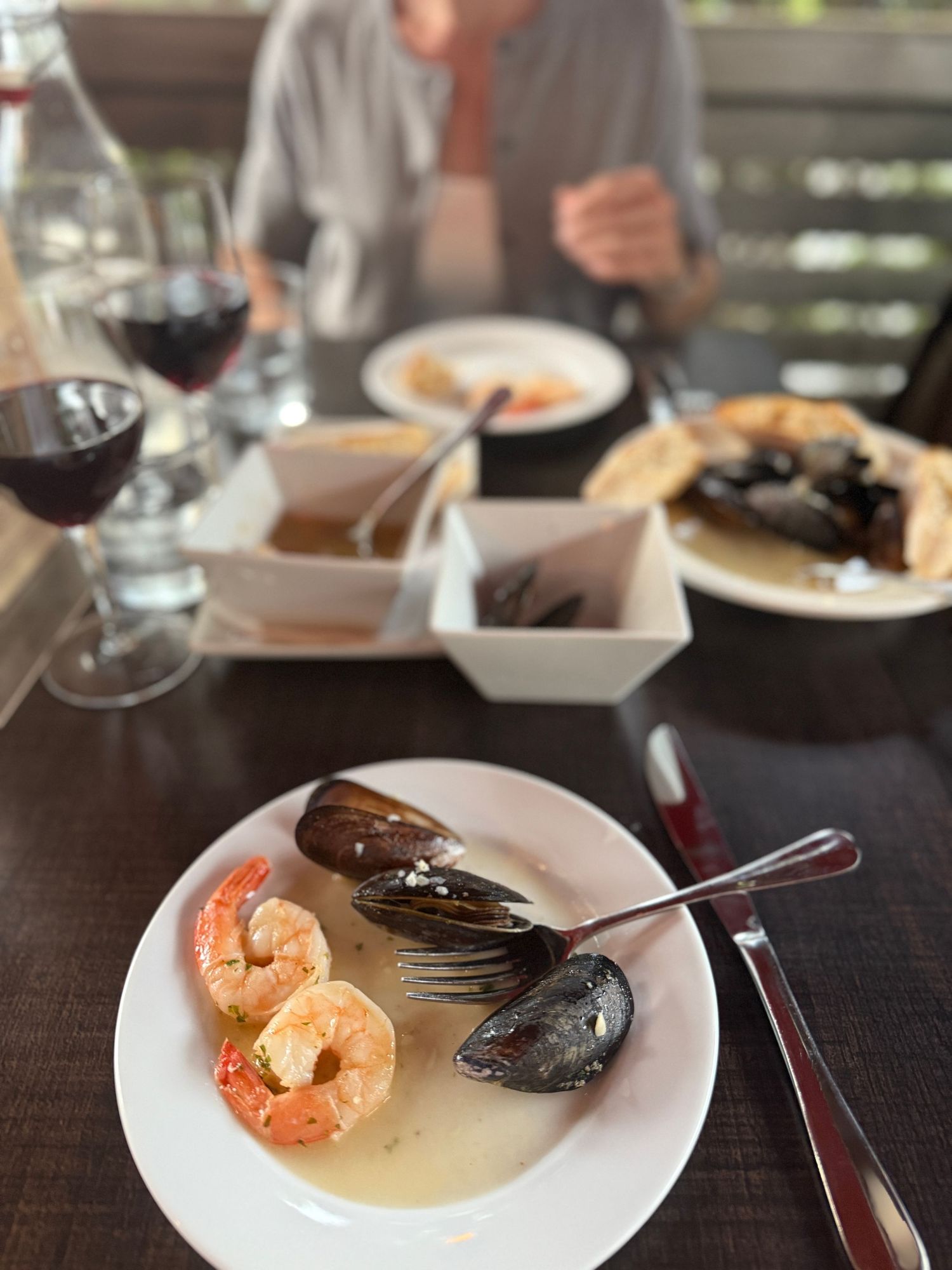 A plate of seafood featuring shrimp and mussels in broth served alongside glasses of red wine at a restaurant table.