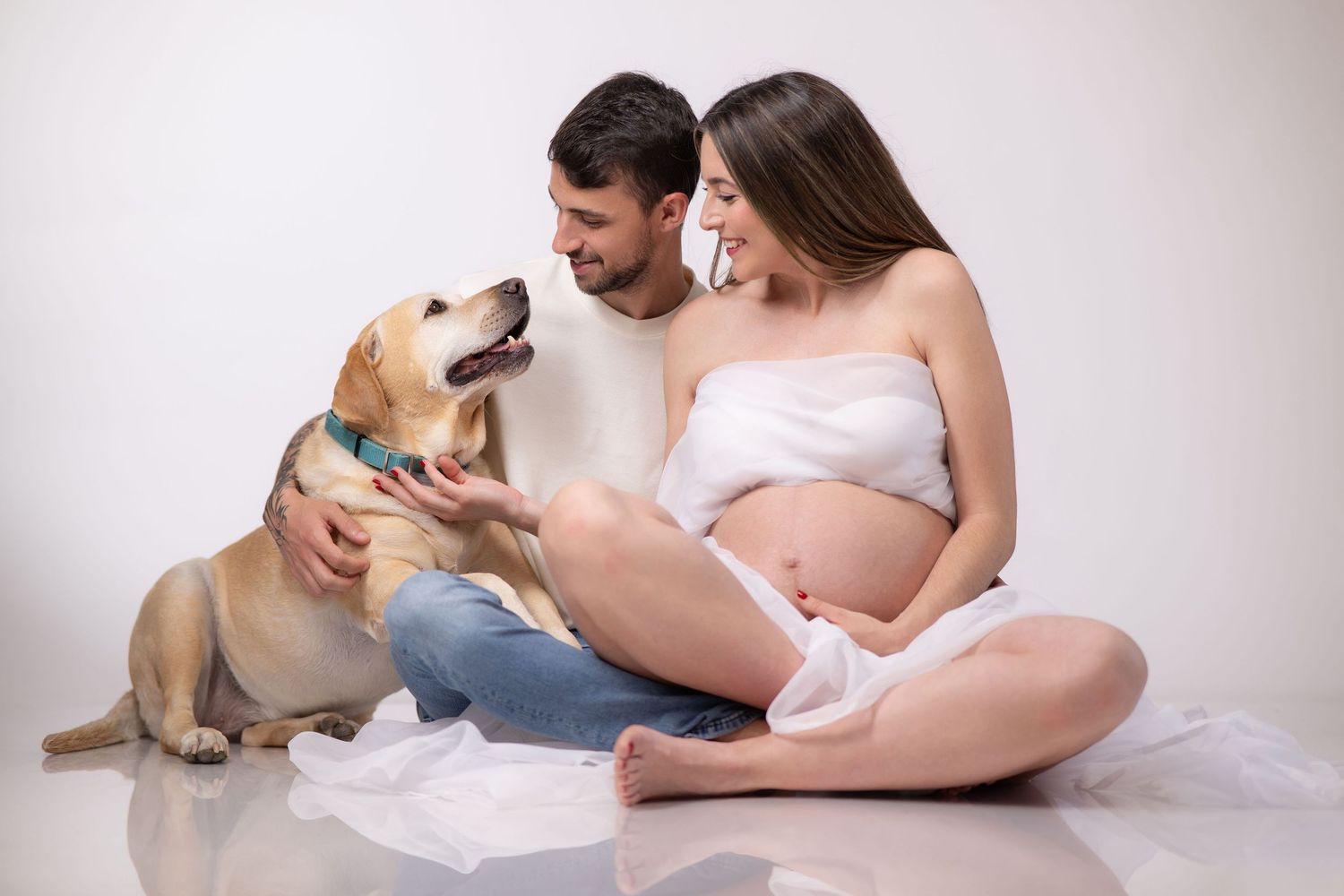 A pregnant woman in white clothing embraces her partner on a sunlit porch during a maternity photoshoot.