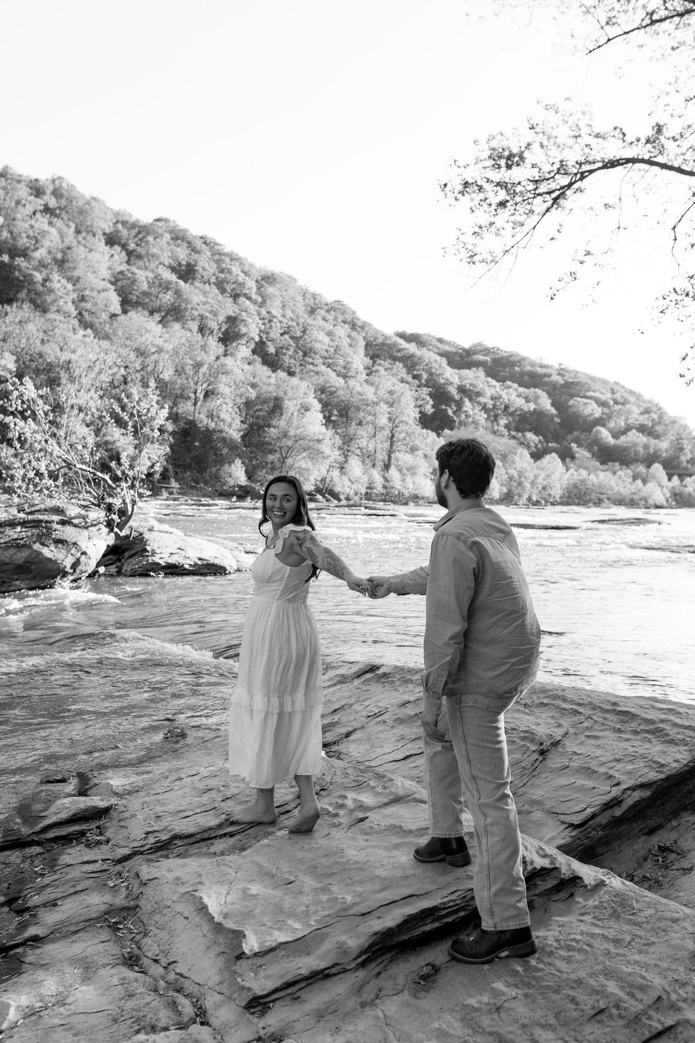 Black and white photo of couple holding hands on rocky riverbank with forest behind.