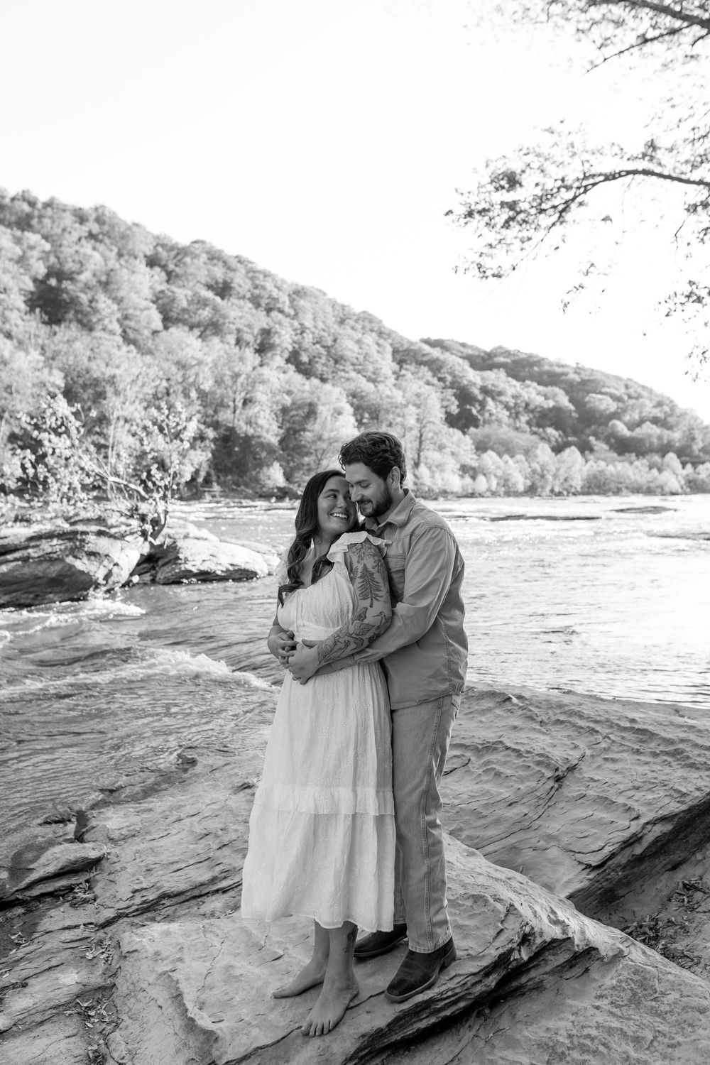 Black and white photo of a couple embracing on rocky riverbank with forested hills in background.