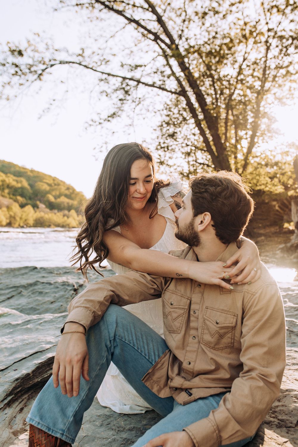 Couple sitting on rocks by a river, woman in white dress lovingly touching mans face in golden sunlight.