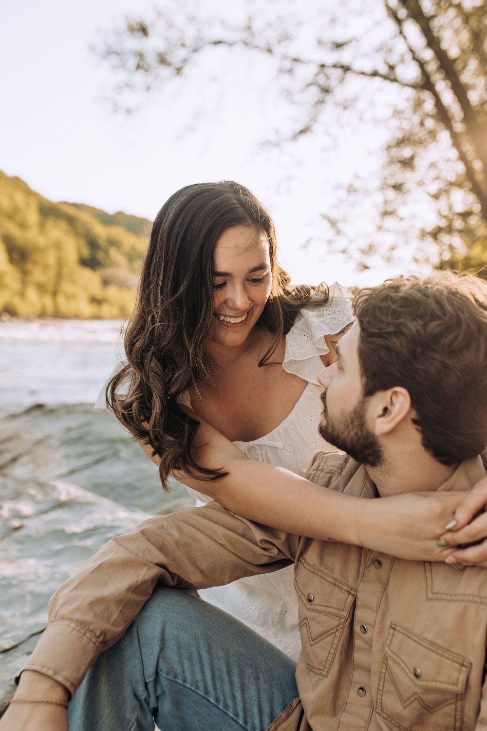 Laughing couple sitting together on rocky riverbank, sharing a joyful moment in warm golden afternoon light.