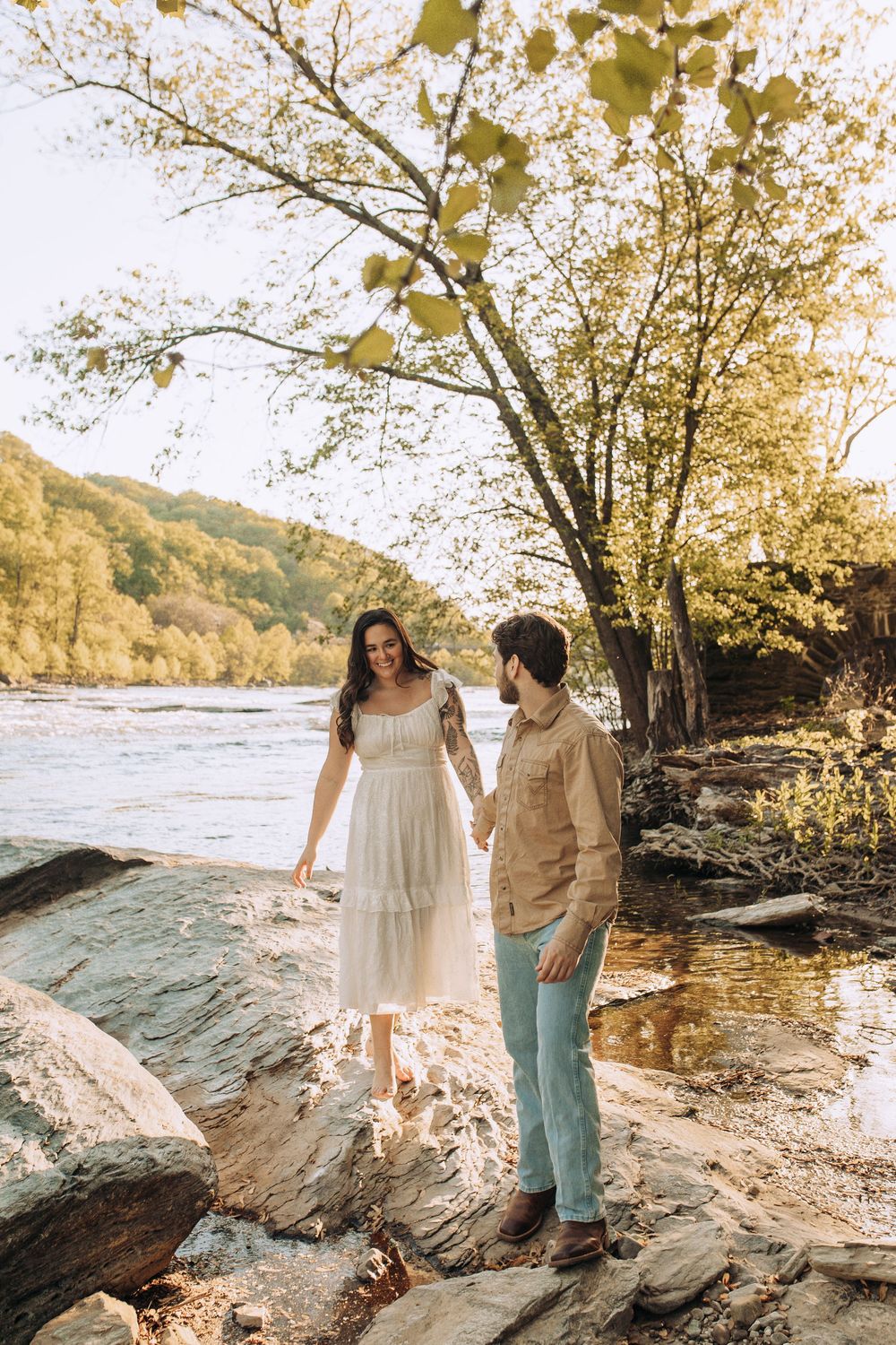 Couple holding hands on rocky riverbank, woman in white dress smiling at partner.