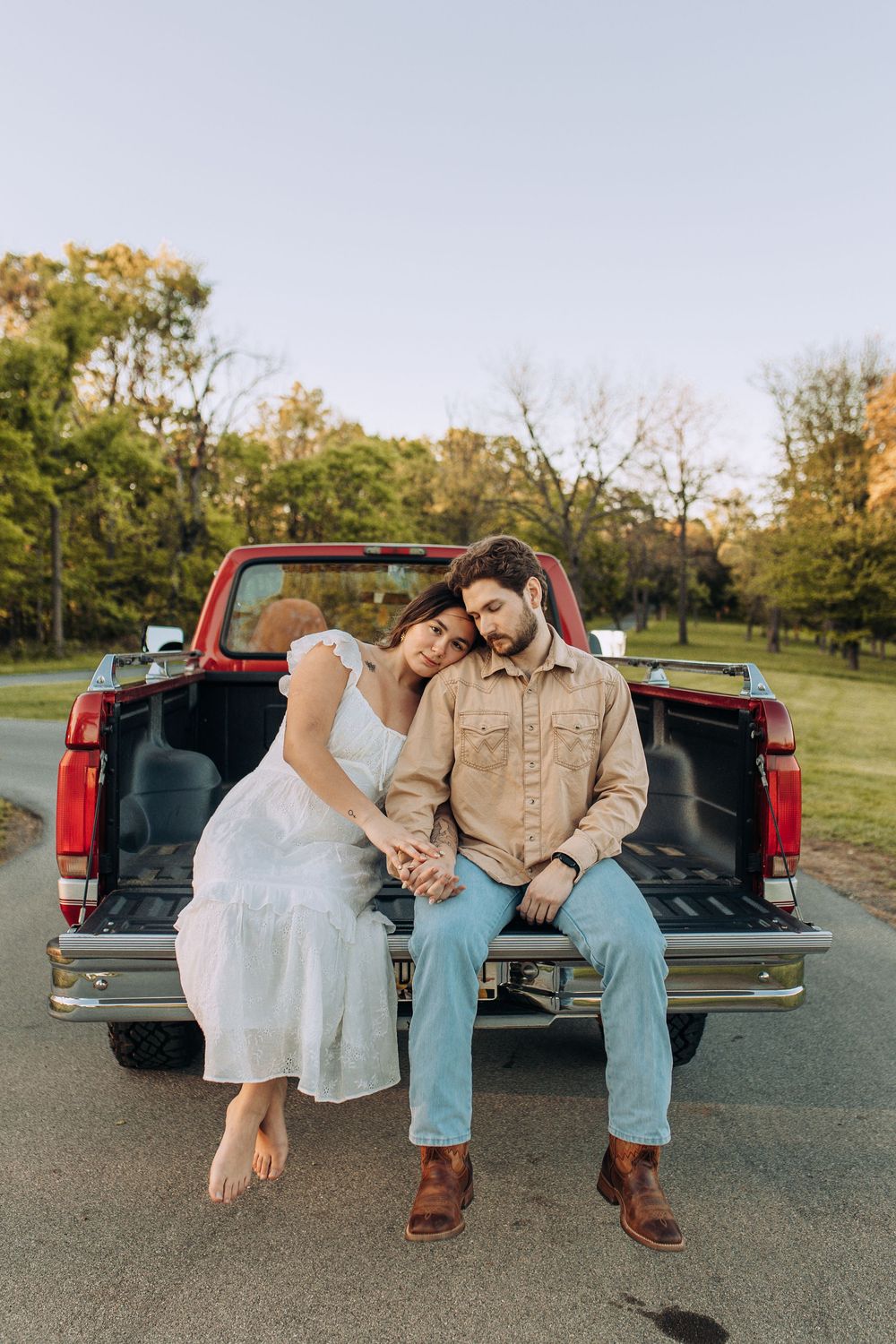 Couple sitting together on a red pickup truck tailgate holding hands at dusk.