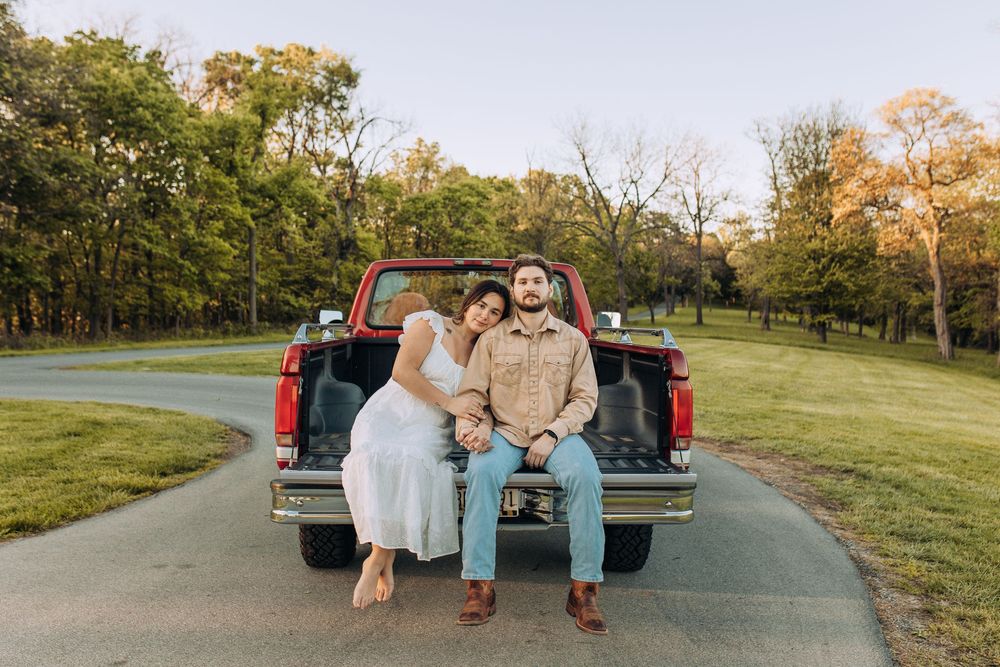 Couple sitting on a red pickup truck tailgate smiling in a park at golden hour.
