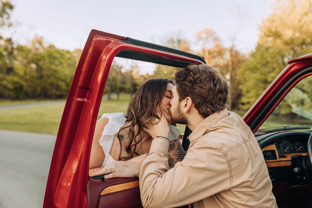 A couple shares a tender kiss leaning through the open door of a vintage red pickup truck surrounded by green trees.