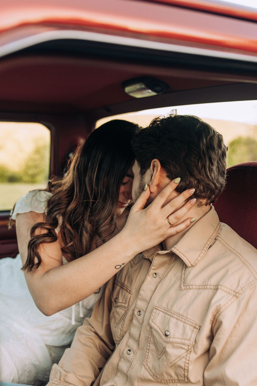 A woman in a white dress pulls a mans face close for a kiss inside the red interior of a vintage pickup truck.