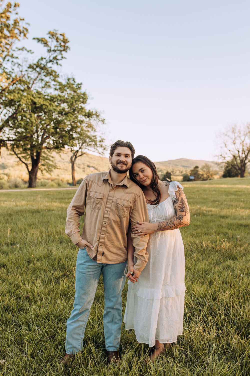 Couple posing together in a sunny green field, woman in white dress leaning on mans shoulder.