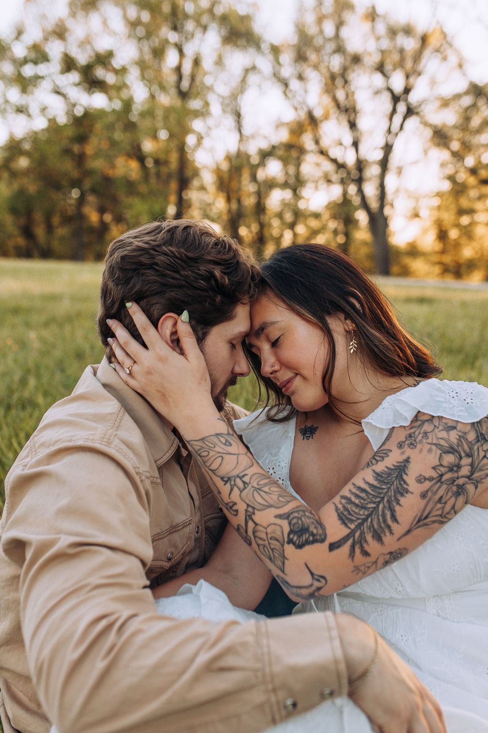Romantic couple touching foreheads and smiling in a sunlit park, woman with tattoos in white dress holding mans face.