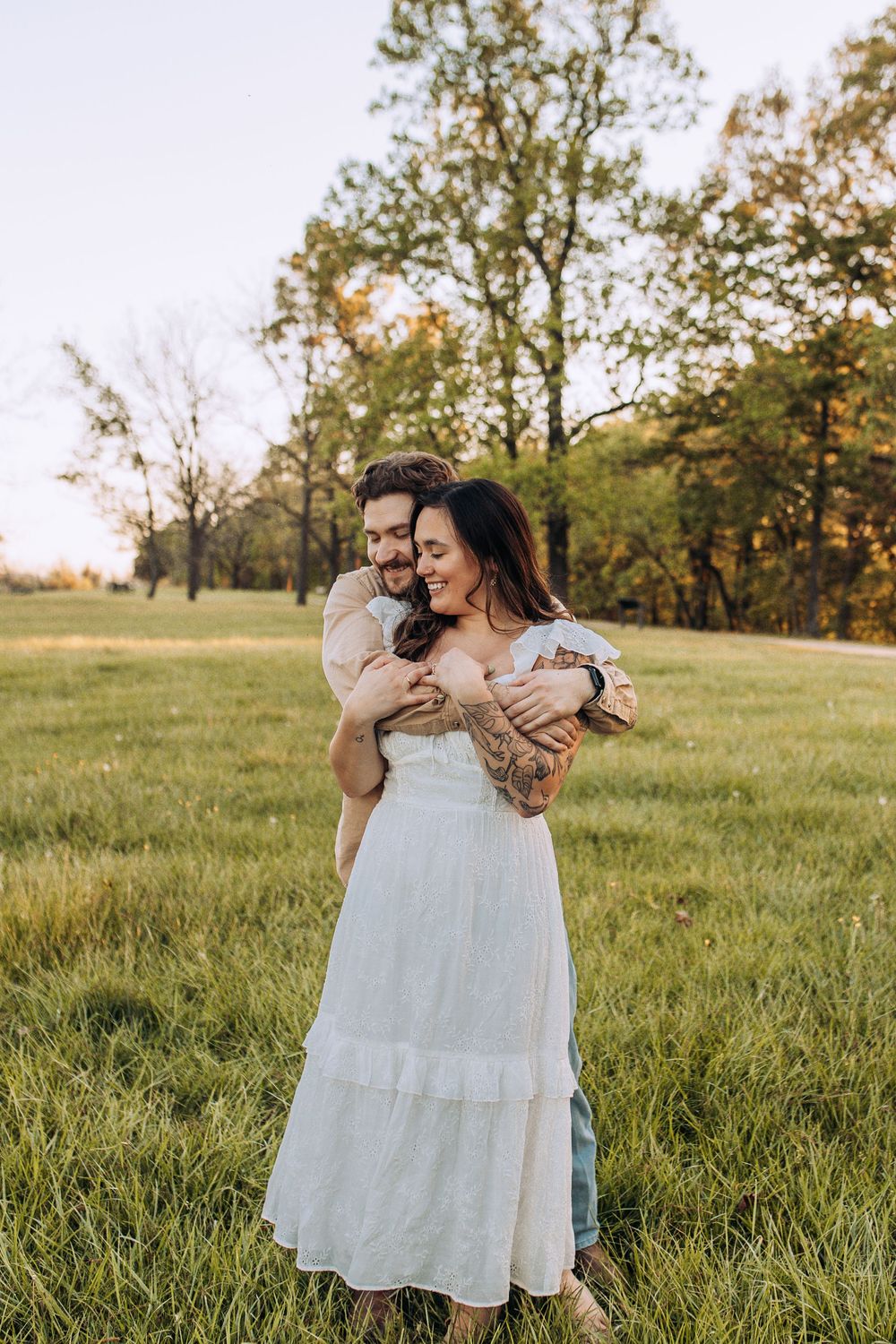 Man hugging woman from behind in an open grassy field at sunset, both smiling and wearing casual outdoor attire.