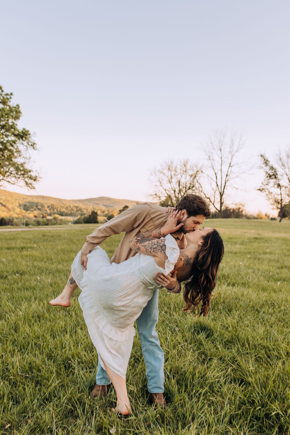 Man dipping woman for a kiss in a lush green field at sunset, with rolling hills and trees in the background.