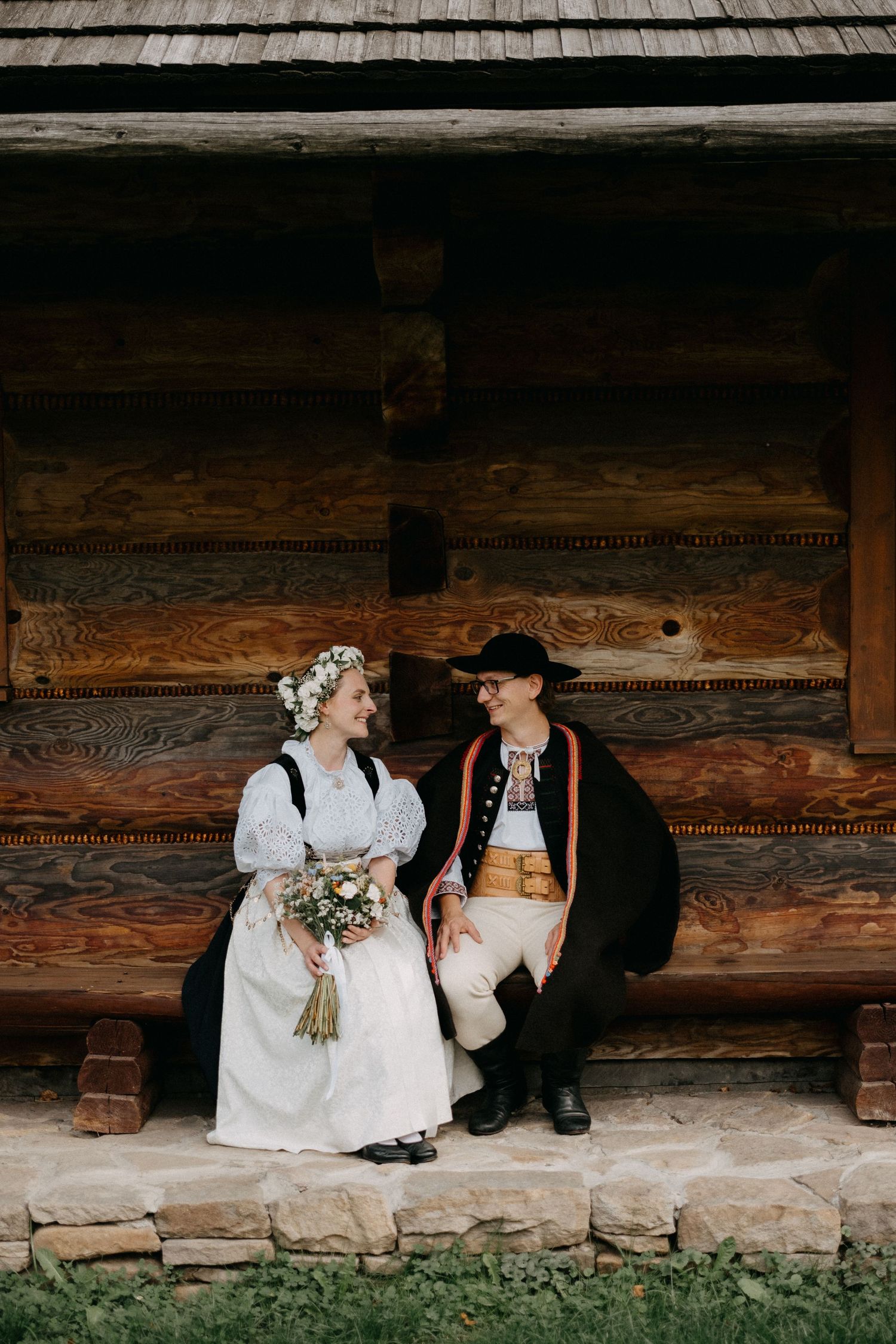 A couple in traditional folk costumes sits together on a rustic log cabin porch with wooden shingles overhead.