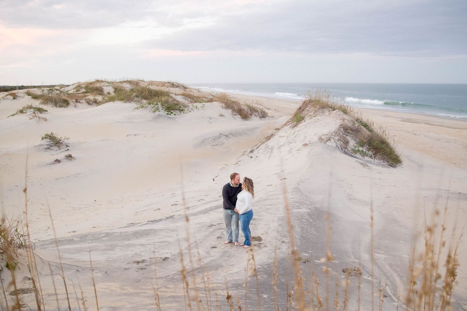 A person in a pink dress stands on sandy dunes at sunset with cloudy skies in the background.
