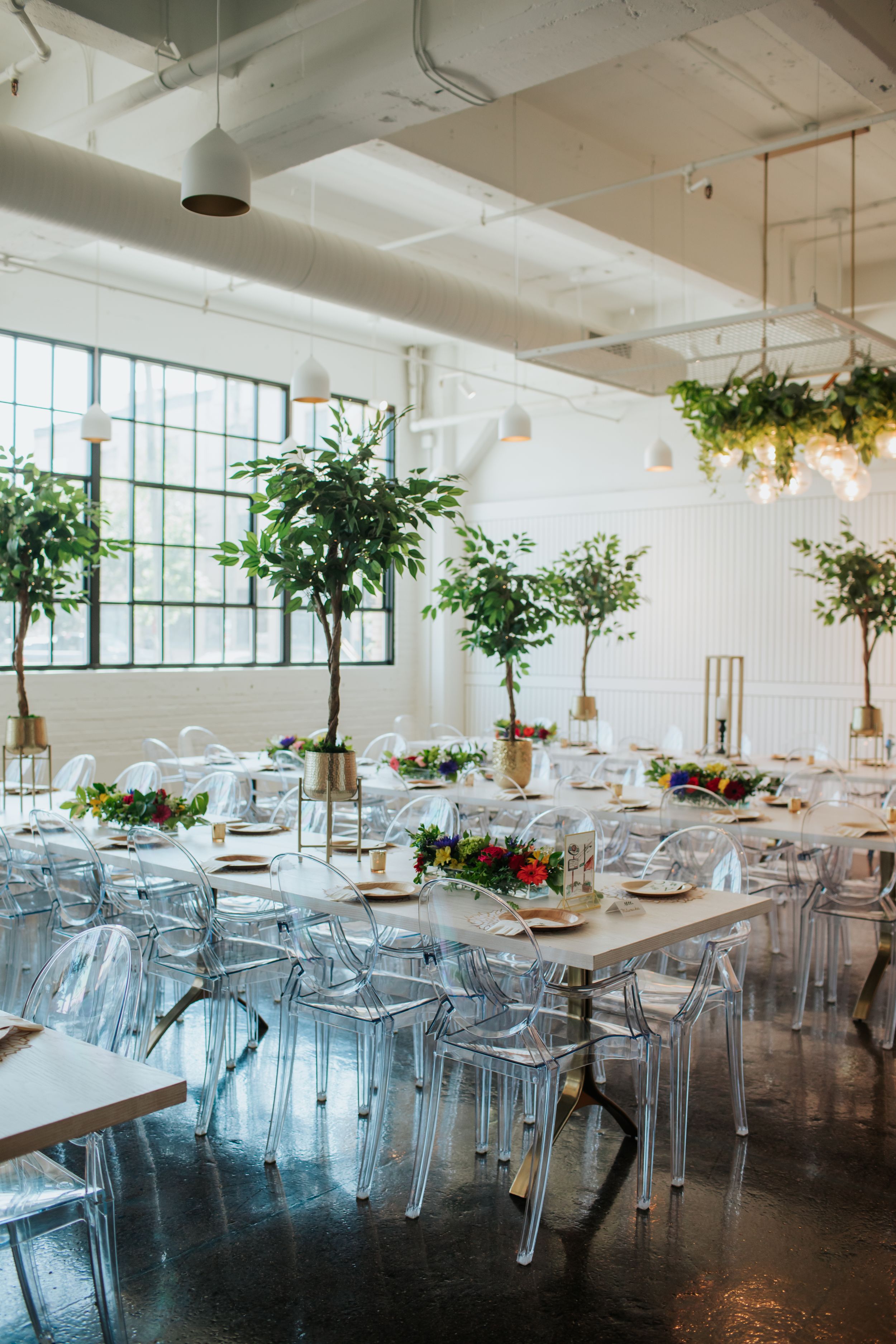 Modern event space with white tables, transparent chairs, and hanging plants creating an airy, industrial-chic atmosphere.
