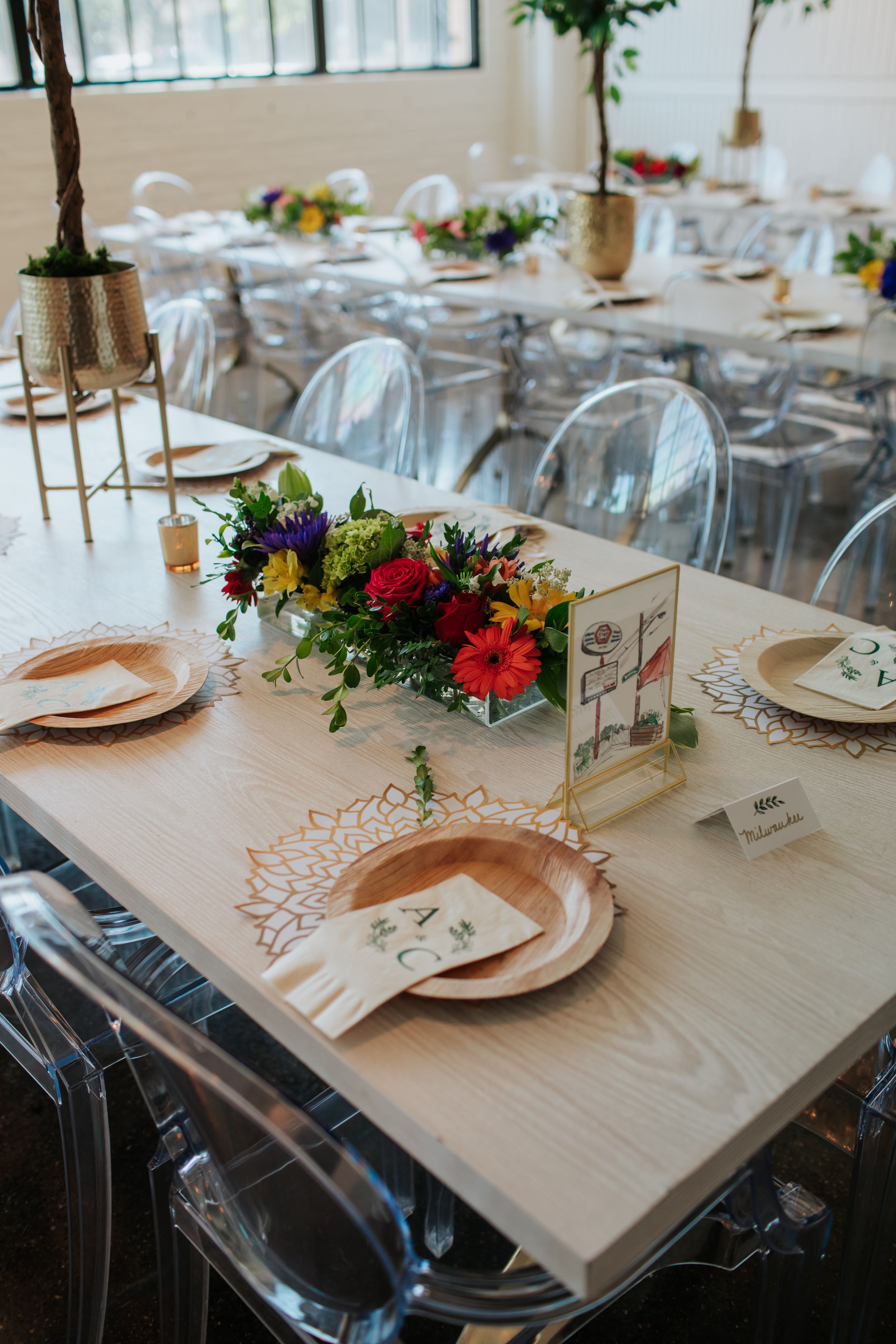 Elegant wedding reception table setting with clear ghost chairs, pink plates, and colorful floral centerpieces.