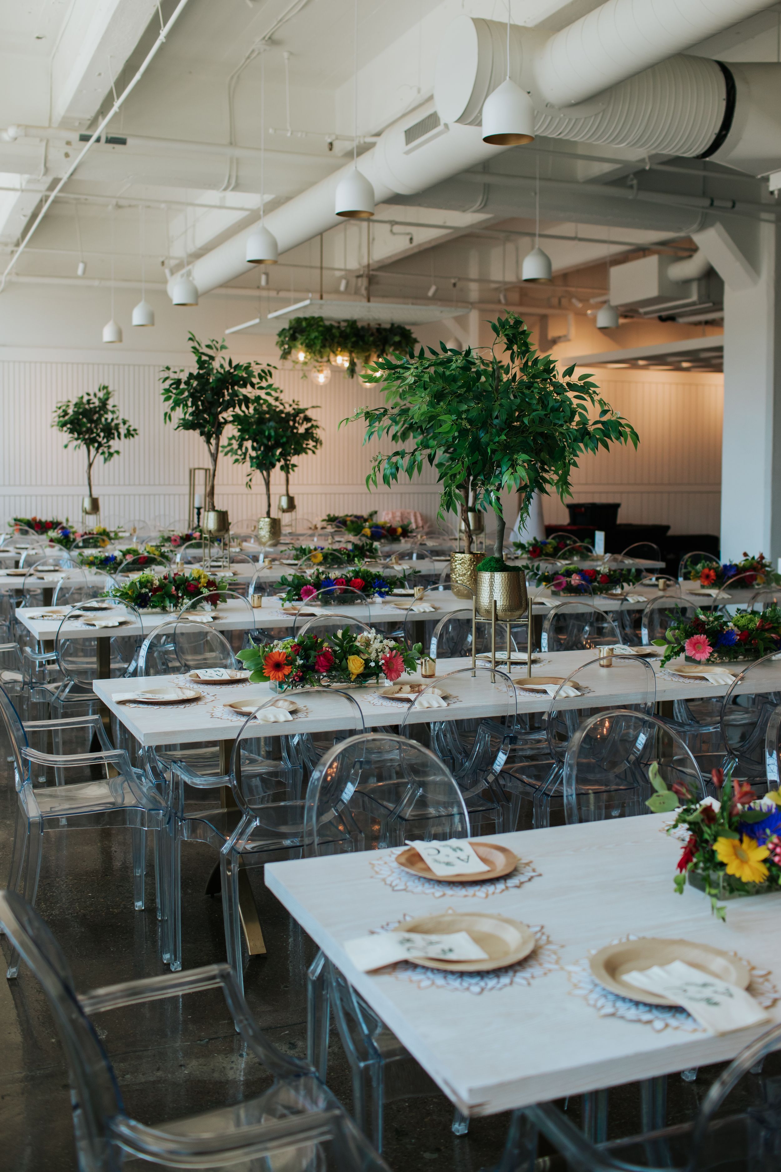 White reception tables arranged with clear ghost chairs and potted trees in a bright industrial venue space.