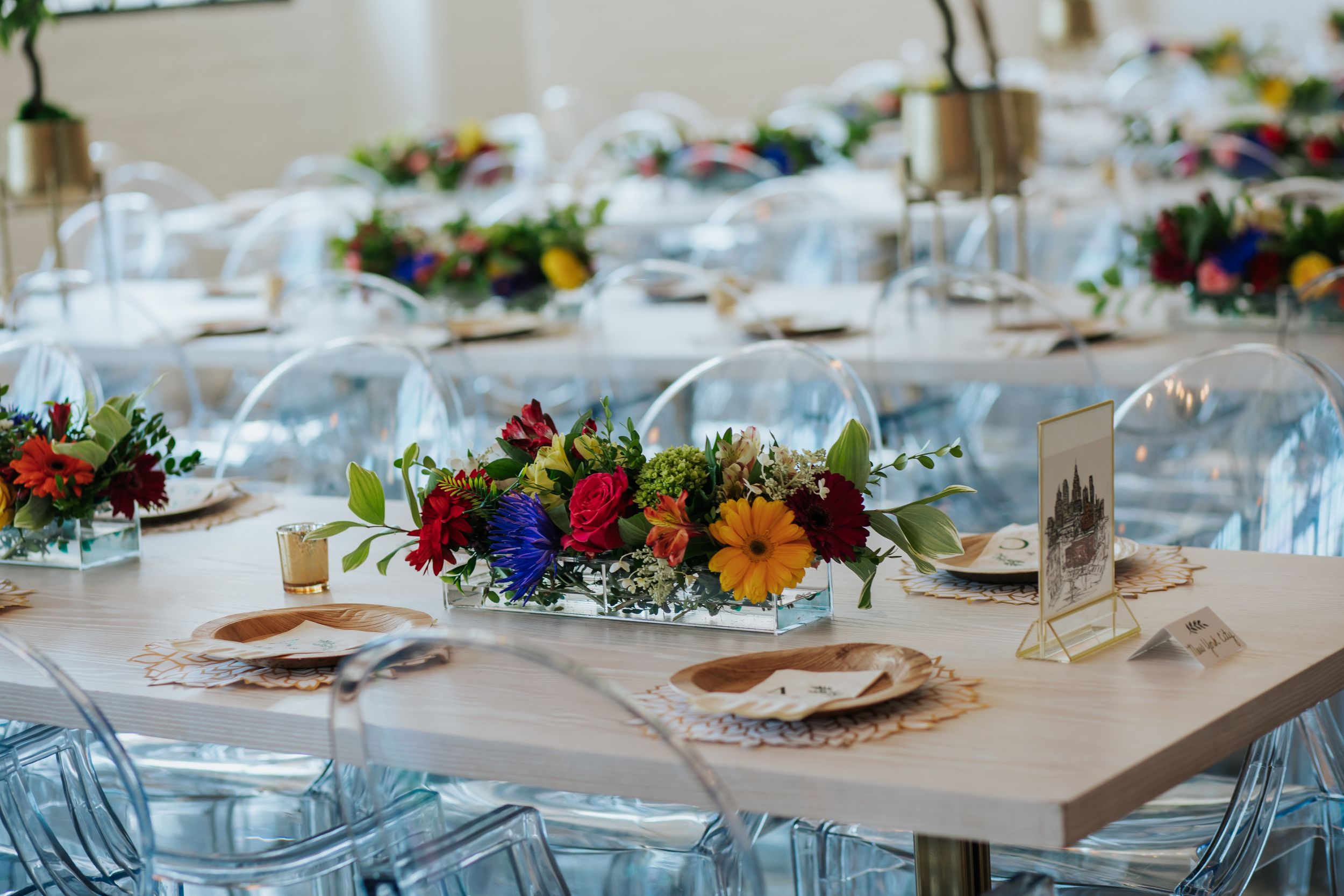 Elegant wedding reception table with colorful floral centerpieces and clear acrylic chairs at an indoor venue.