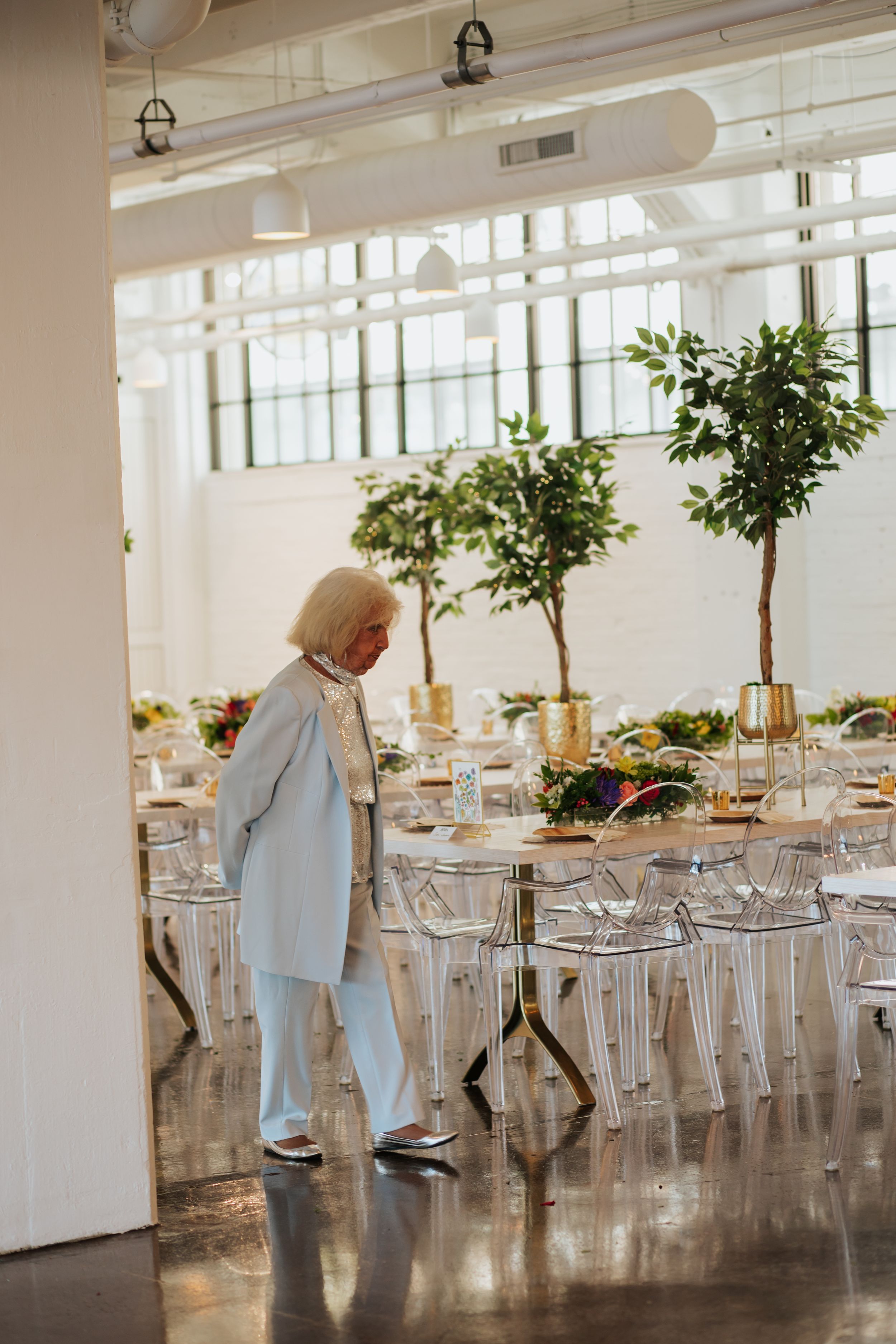 Bright industrial event space featuring modern white tables, transparent chairs and decorative potted trees.