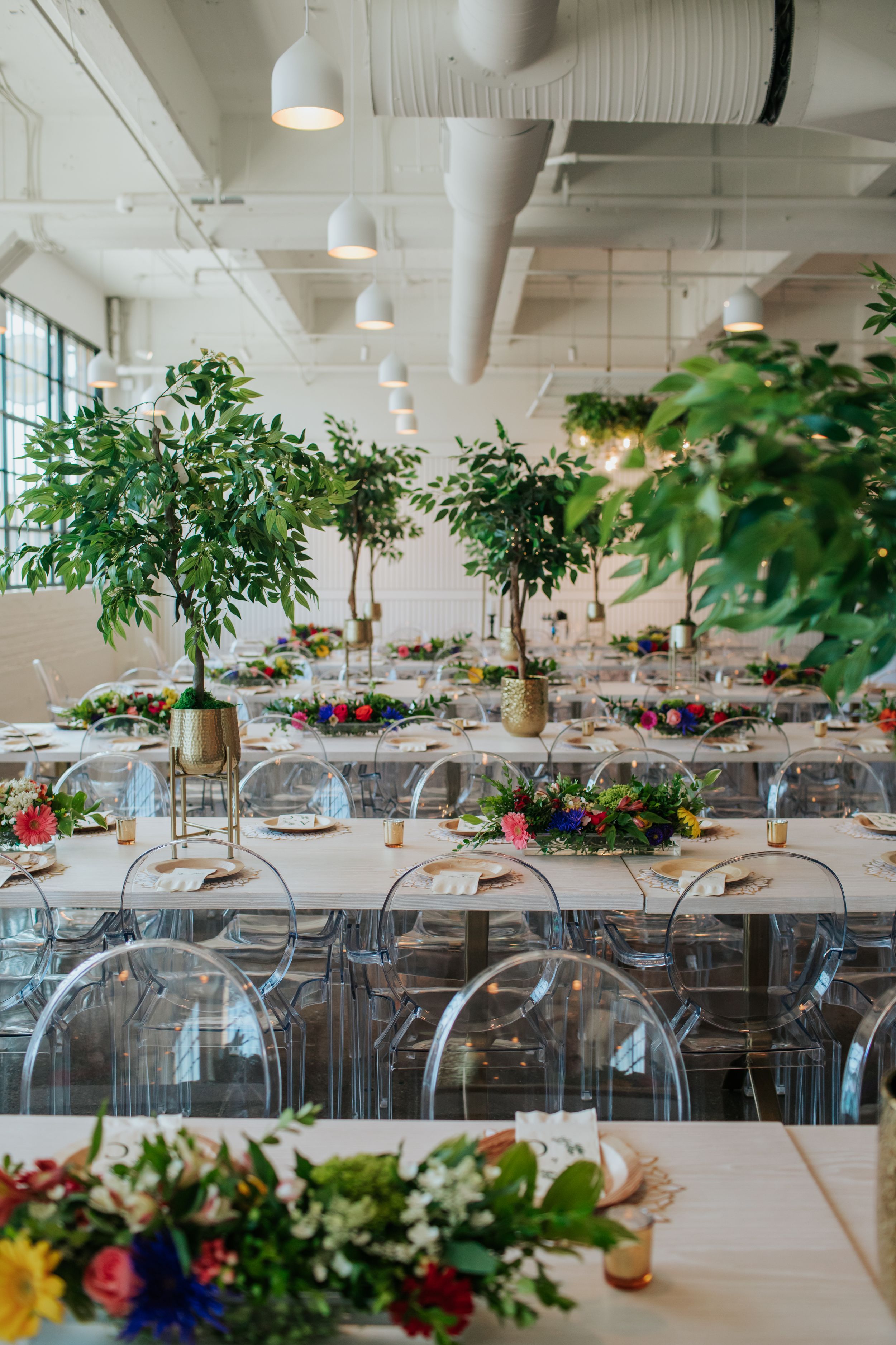 Modern wedding reception venue with clear chairs, potted trees and floral table arrangements.