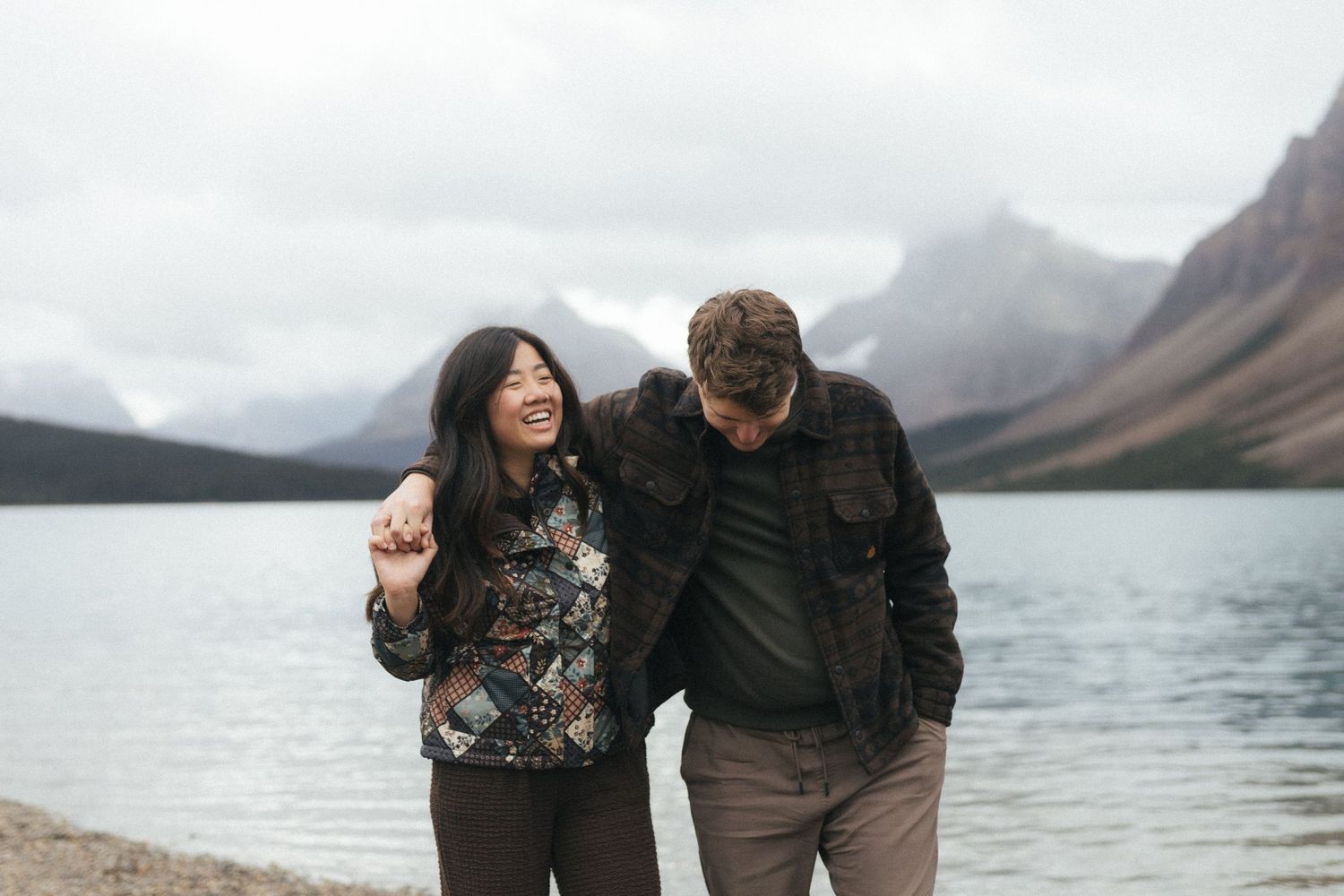 couple in fall jackets laugh and hug on the edge of a mountain lake in Kananaskis