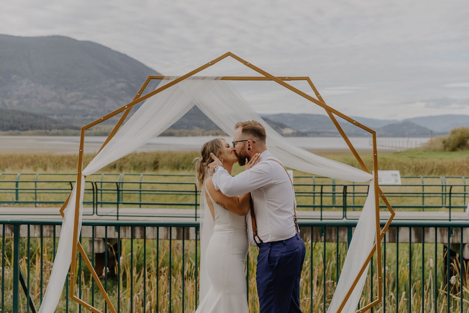 Bride and groom sharing first kiss under geometric gold arch with mountain lake backdrop at outdoor ceremony.