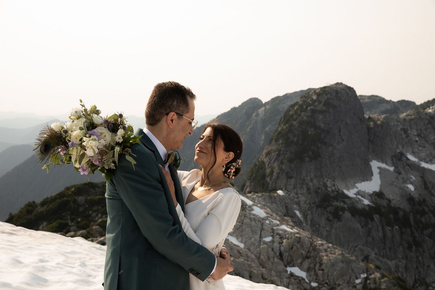 Bride and groom embrace on snowy mountain peak with dramatic rocky landscape behind them.
