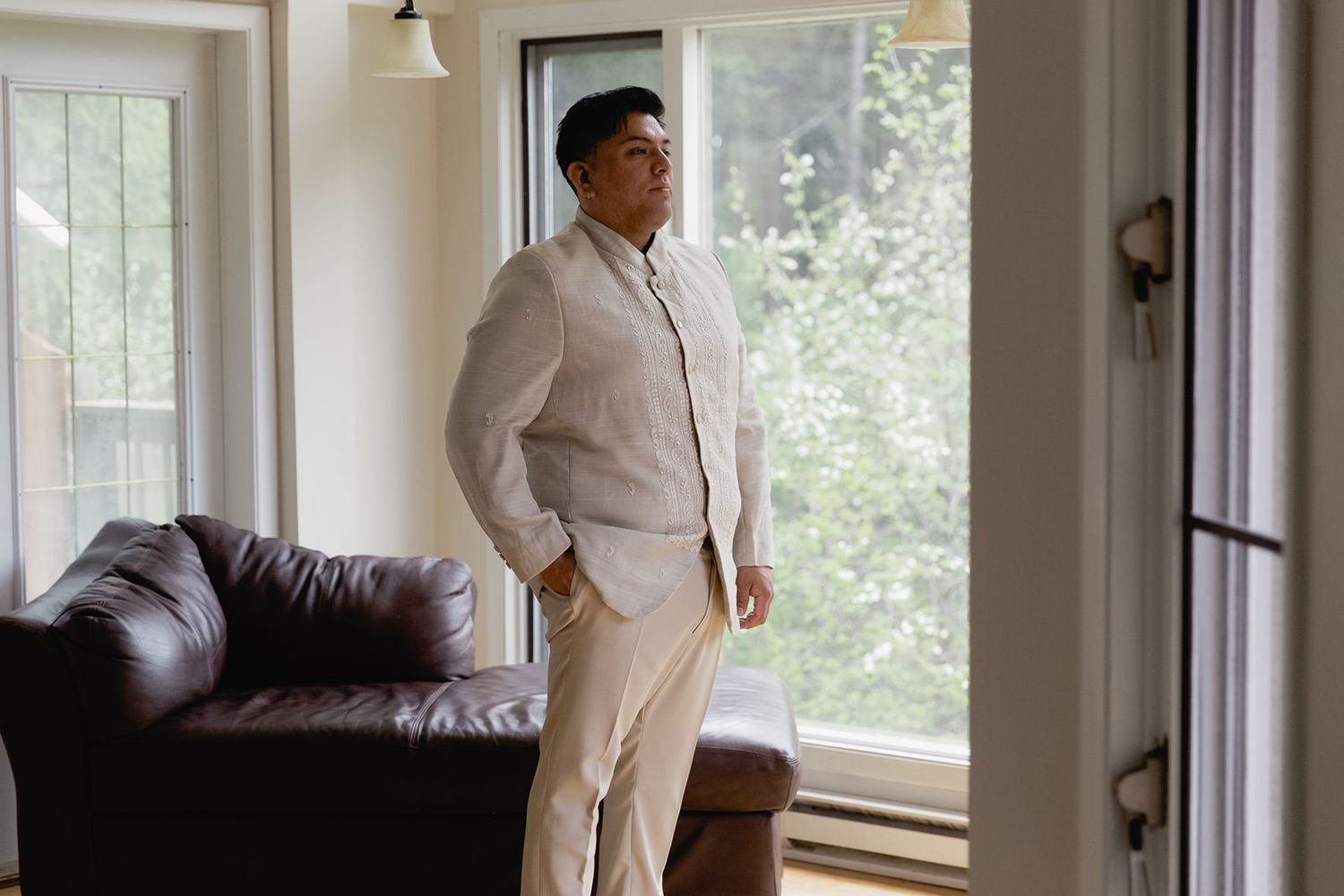 Groom in a cream linen suit stands confidently near large windows in a bright modern room before the wedding.