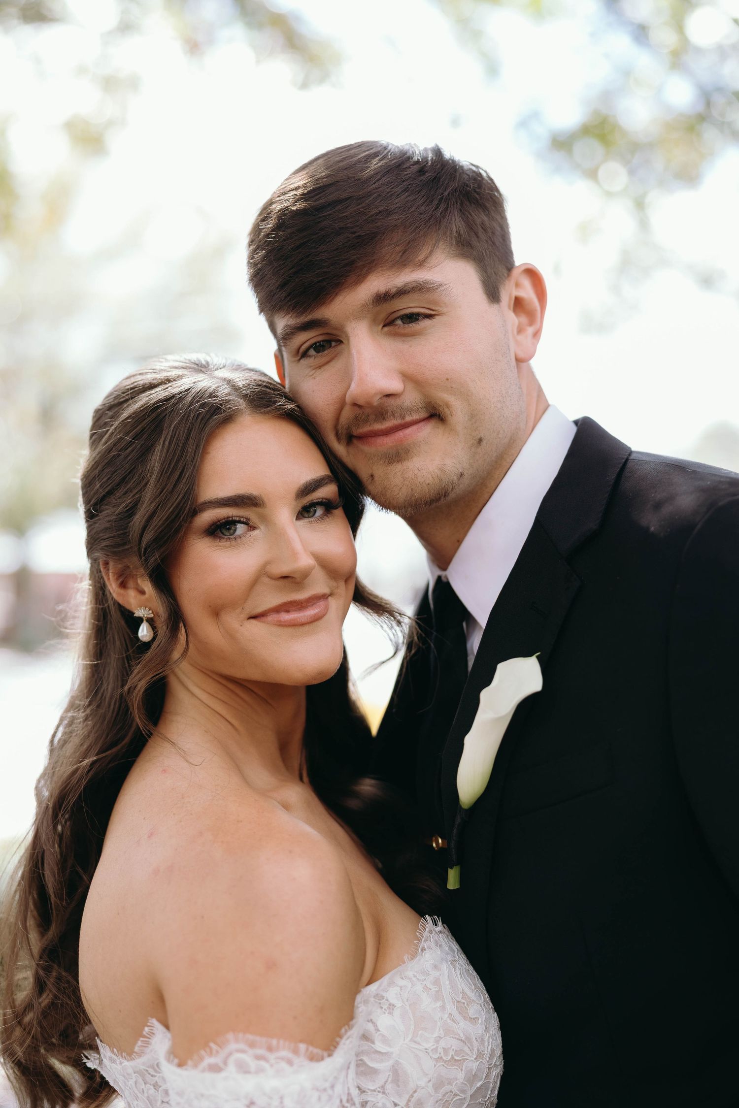 A close-up wedding portrait of a couple in formal attire with a white boutonniere and strapless lace dress.