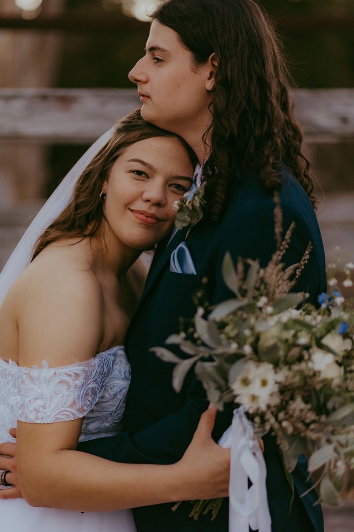 Bride in off-shoulder lace gown leans on groom in navy suit holding a wildflower bouquet at golden hour.