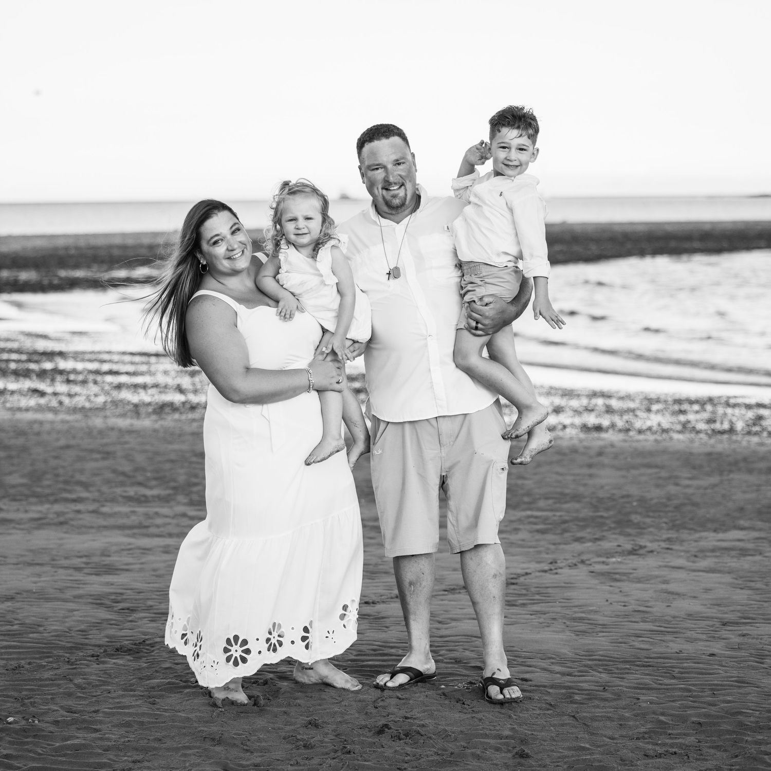 Happy family of four posing on beach in black and white photo, parents holding young children.