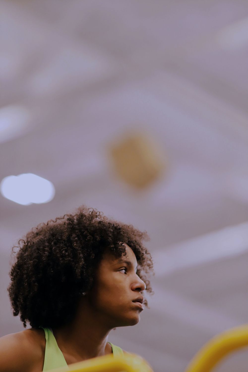 Person with natural curly hair in yellow tank top looking to the side against blurred background.