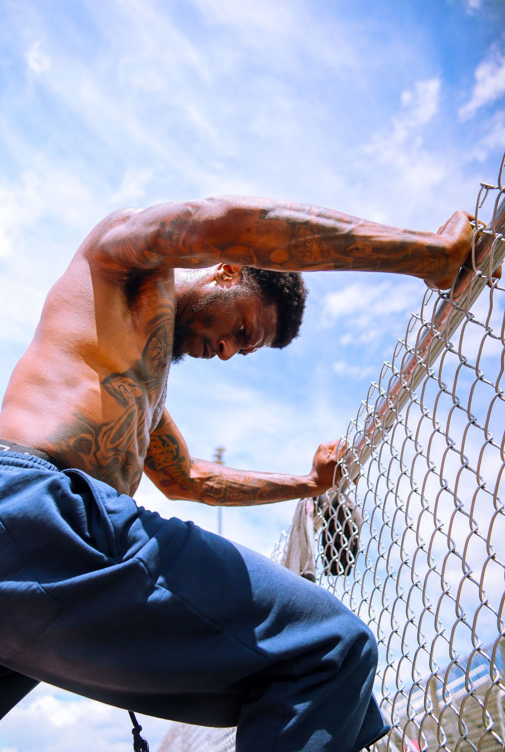 Muscular tattooed shirtless man climbing a chain-link fence against a blue sky backdrop.