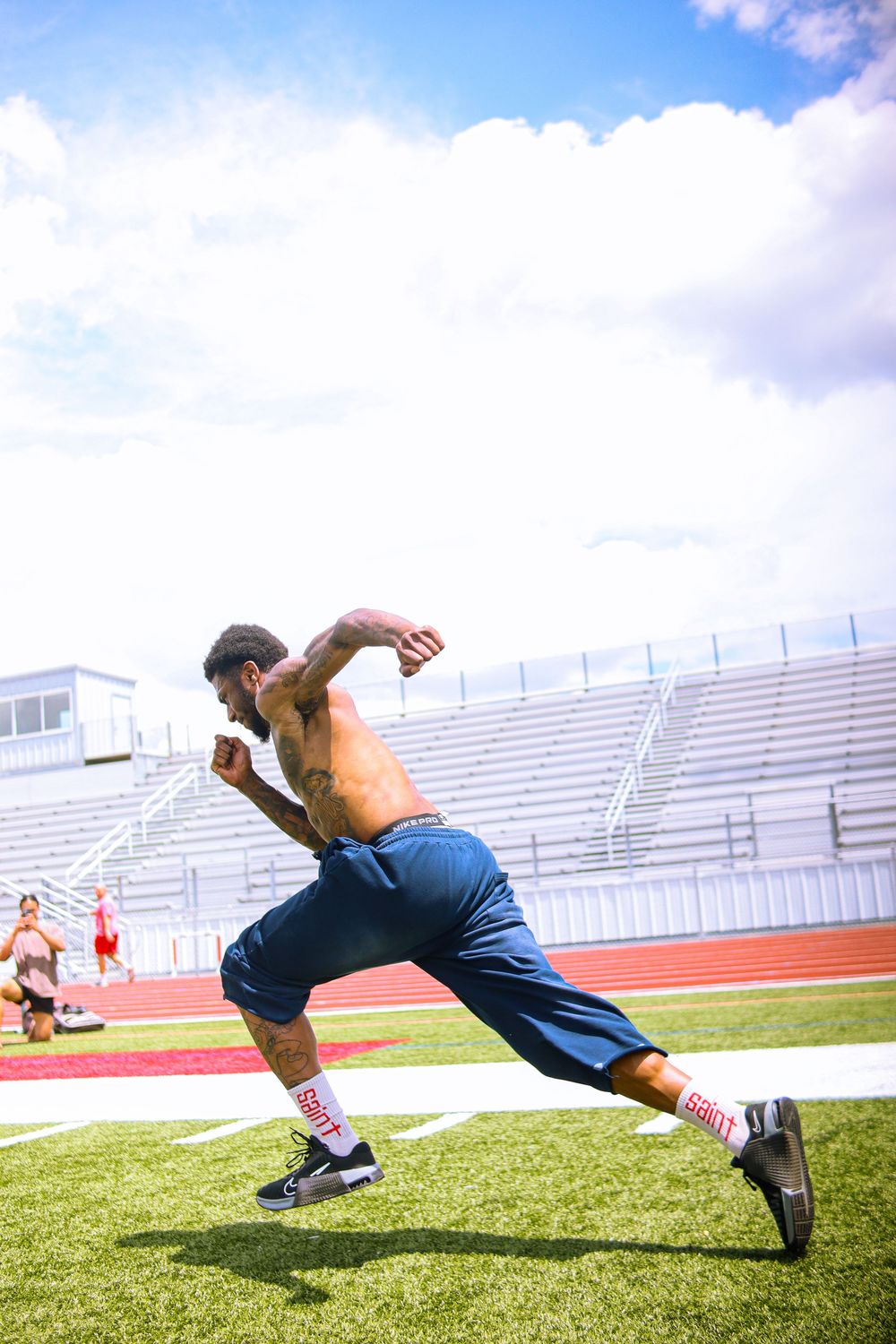 Shirtless athlete sprinting on green turf field at stadium during outdoor football training session.