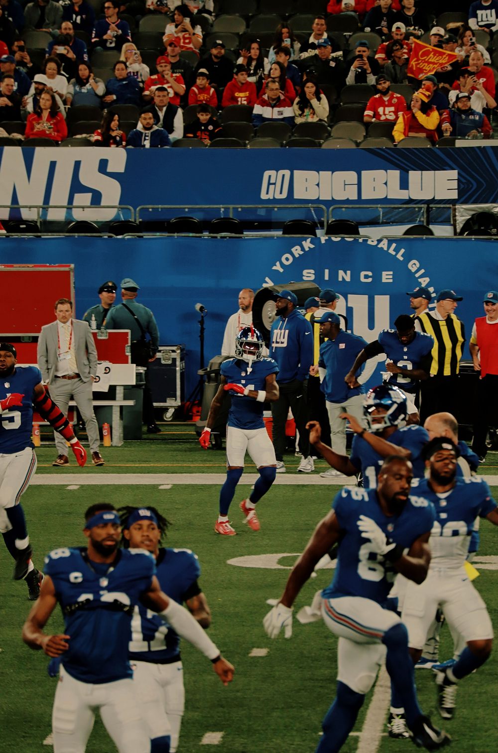 Football players in blue uniforms celebrate on the field during a game with a 'Go Big Blue' banner visible in the background.