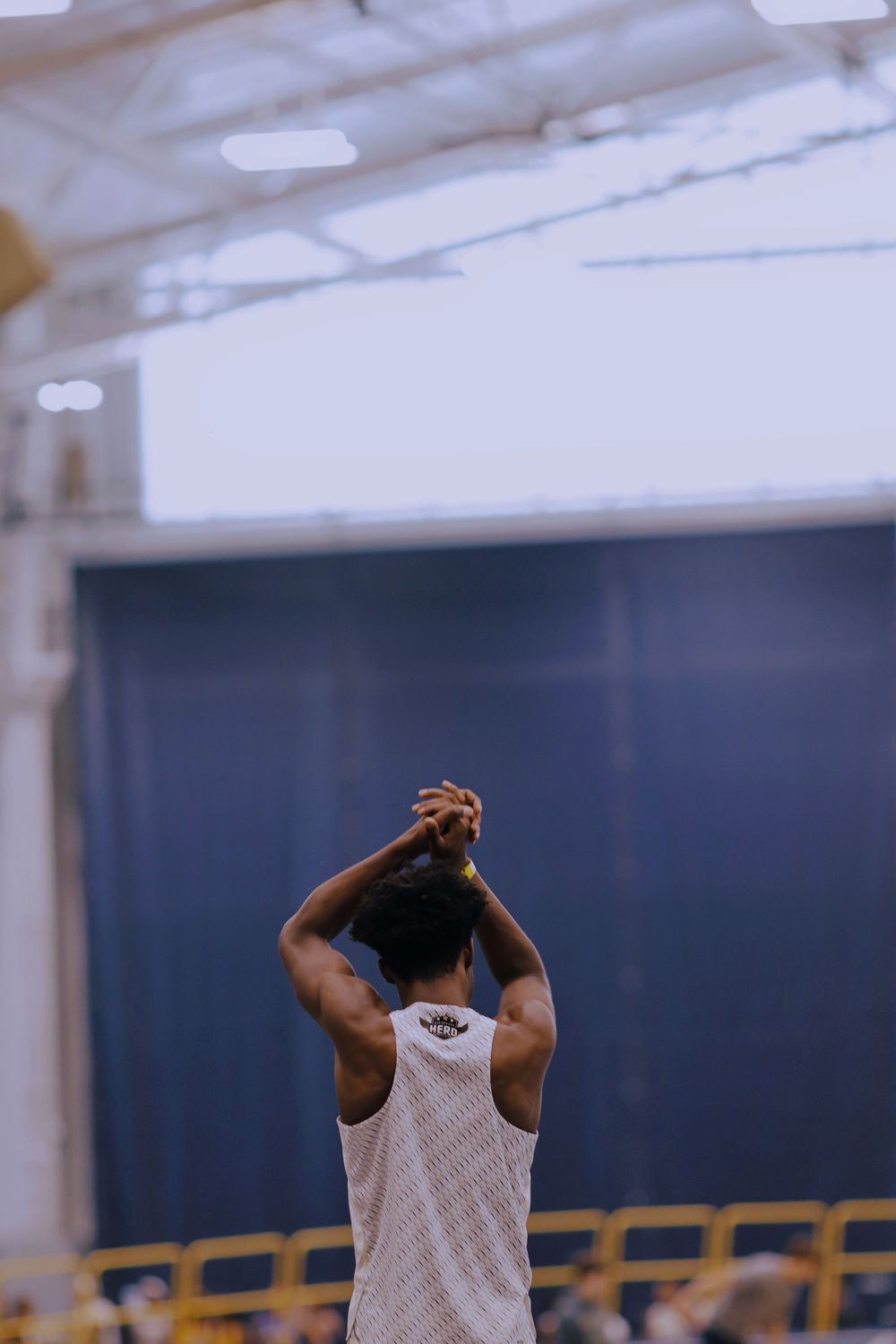 Basketball player in white jersey stretches with hands clasped above head in indoor gymnasium.