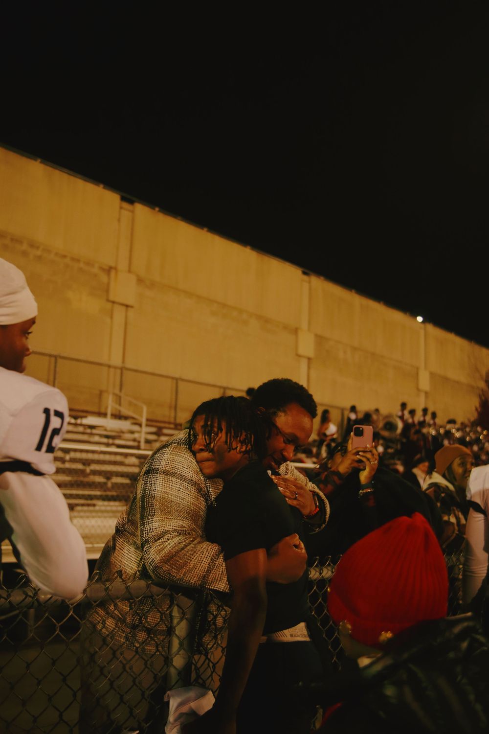 A crowd of spectators watching an event through a chain link fence at dusk.