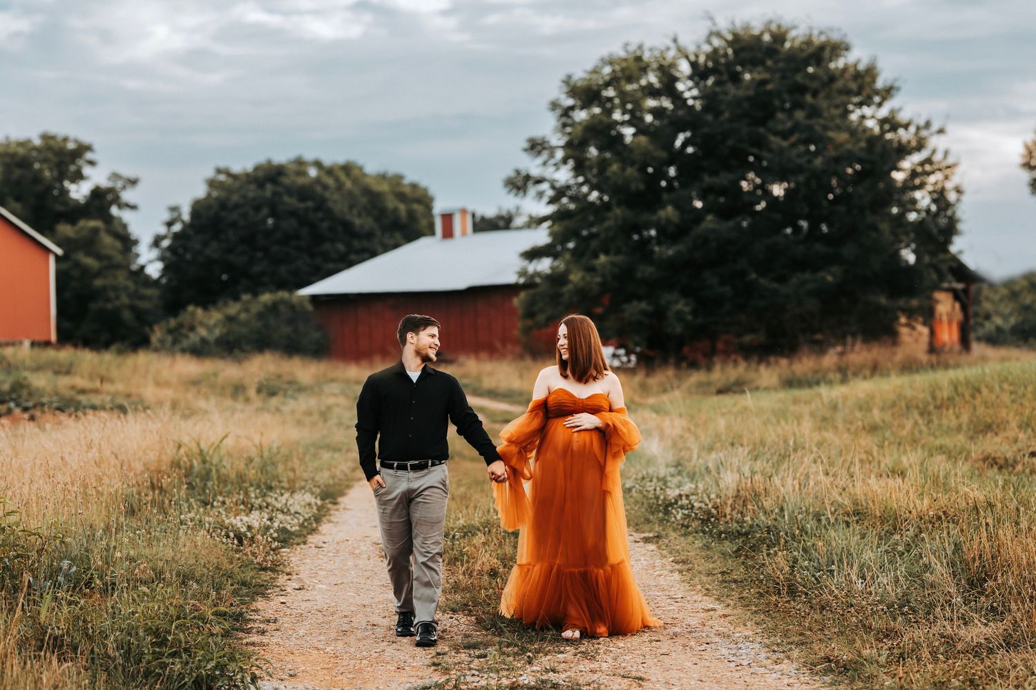 Maternity couple walk together through a rustic countryside field with red barns visible in the background. Maternity Clarksville