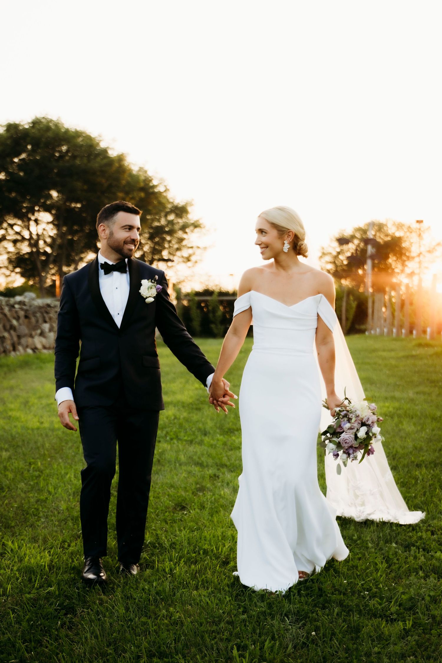 A couple in wedding attire walks through a grassy field at sunset, creating romantic silhouettes against the golden light.