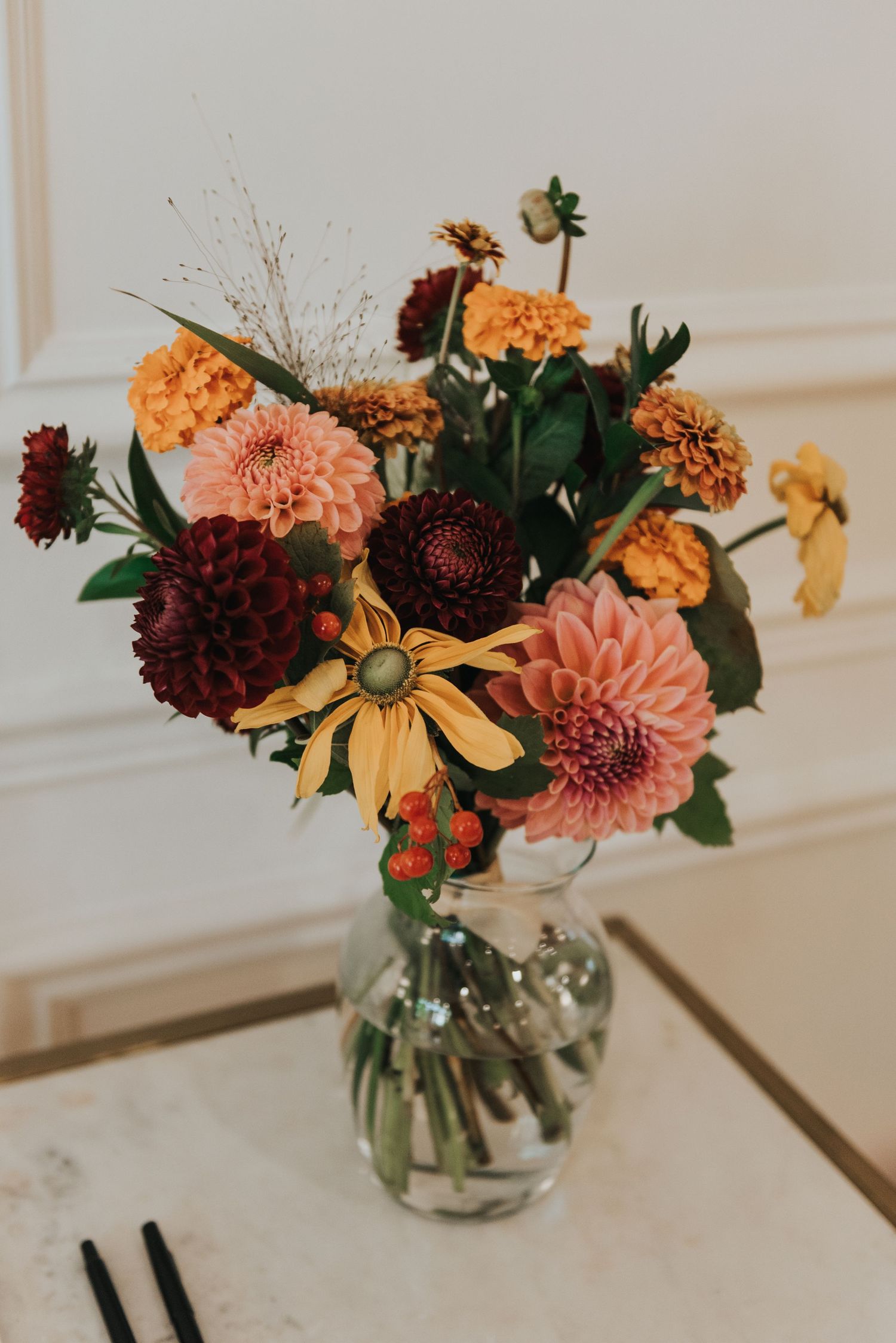 Fall-colored floral arrangement with burgundy and peach dahlias in a glass vase on white marble surface.