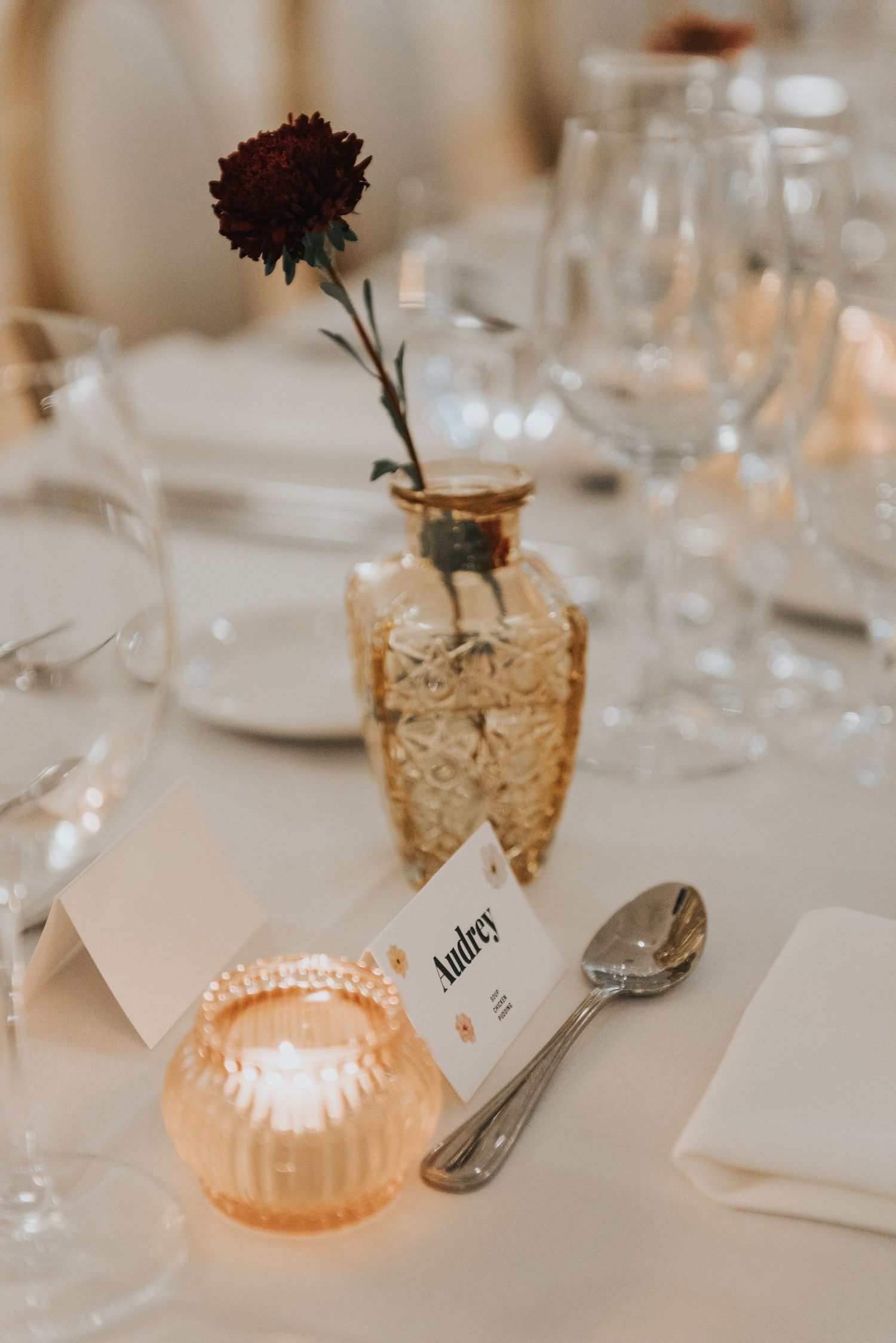 Elegant table setting with dark flower in gold vase, glowing candle holder, and wine glasses on white tablecloth.