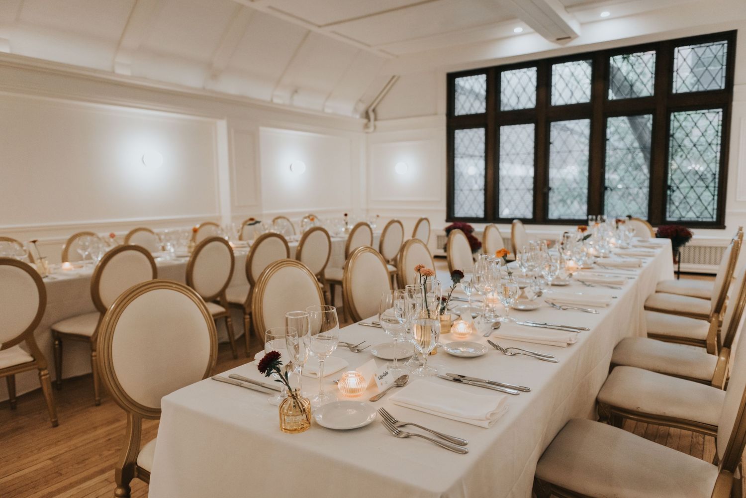 Elegant dining room setup with long white table, cream chairs and natural light streaming through black-framed windows.