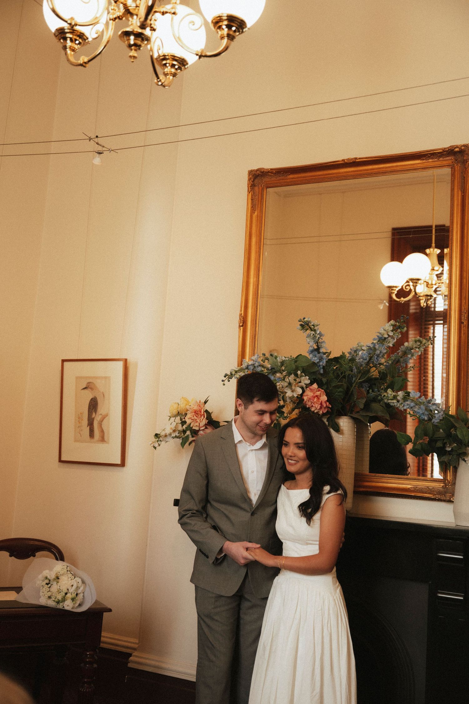 A couple in wedding attire walks through a lush garden path surrounded by blooming flowers and greenery.