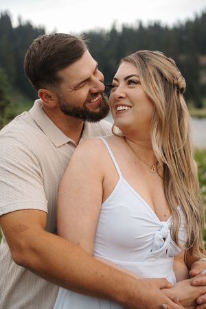 A couple embraces and laughs together during an outdoor photo session with evergreen trees in the background.