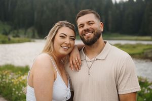 A couple poses together by a mountain lake surrounded by summer wildflowers.