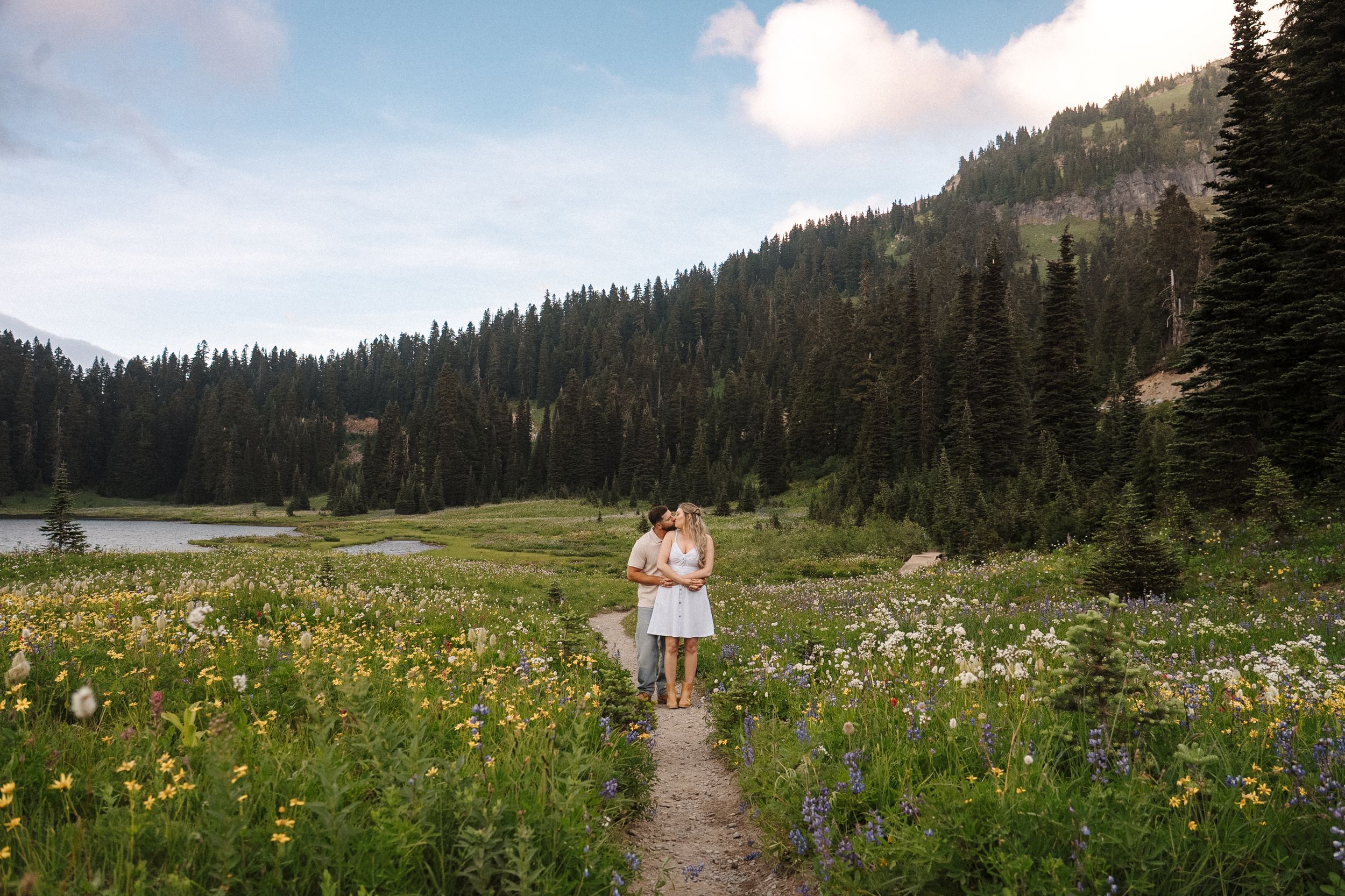 A scenic mountain landscape with wildflowers and a lake surrounded by evergreen forest.