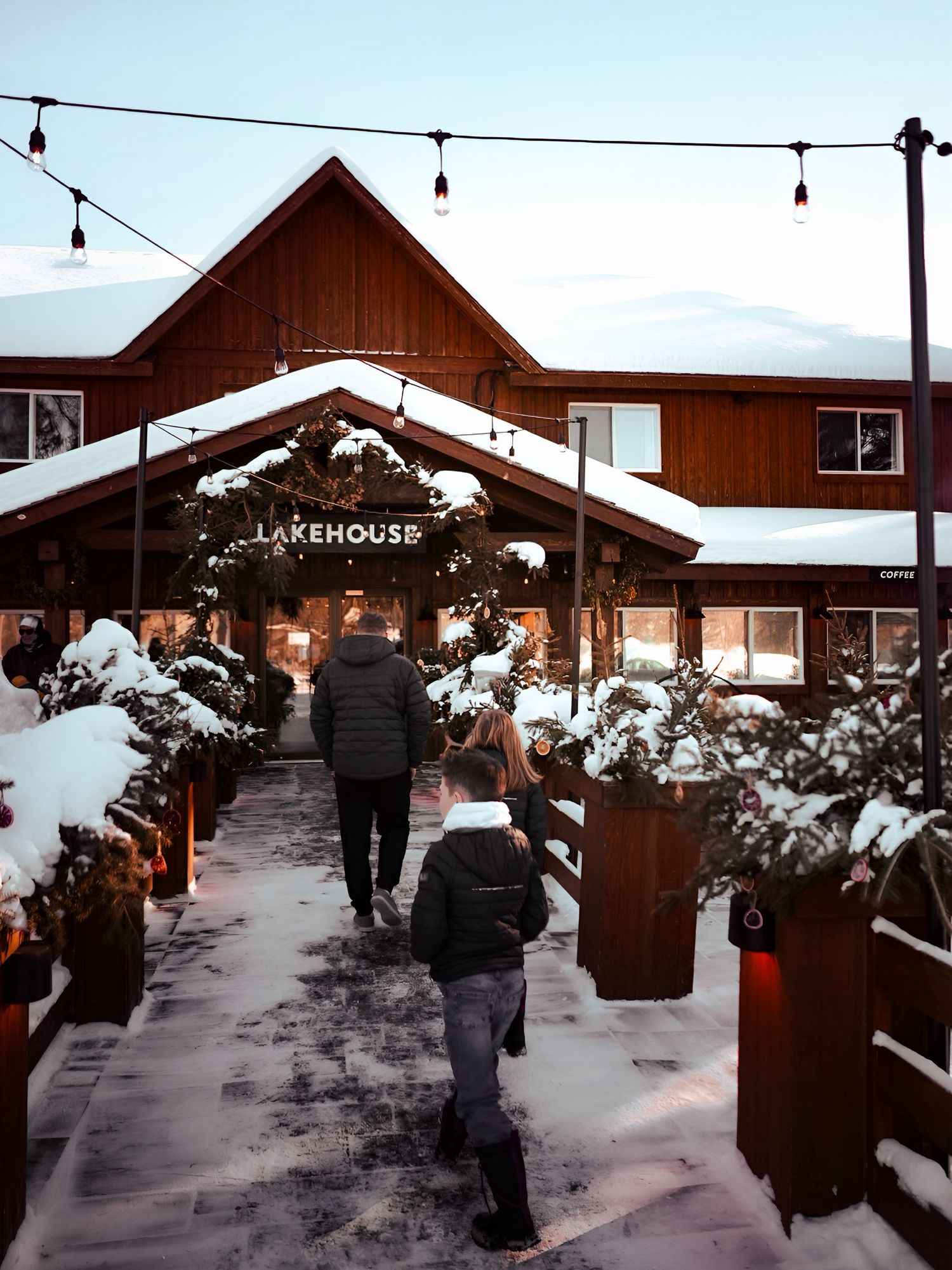 A snowy walkway leads between wooden cabin buildings at a winter resort with snow-covered trees and paths.