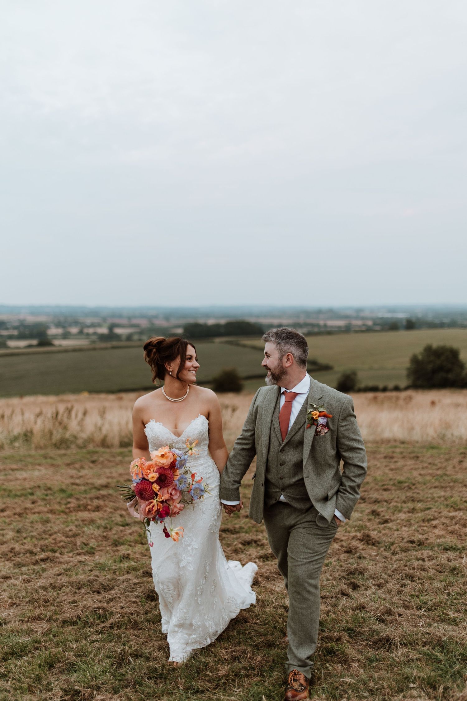A couple in wedding attire walks together on a hilltop overlooking scenic countryside fields at sunset.